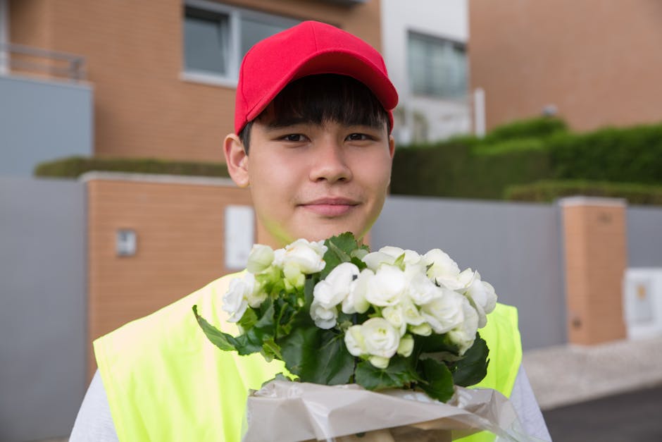 A young man wearing a red cap and a high-visibility yellow vest is holding a bouquet of fresh white flowers wrapped in white paper. The bouquet features delicate, fully bloomed roses and lush green le