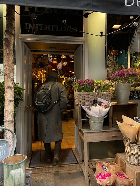 A person dressed in a dark coat and wearing a black backpack is entering 'INTERFLOW' flower shop on Upper Street in Islington, with the shop's interior warmly lit and decorated with hanging lights and