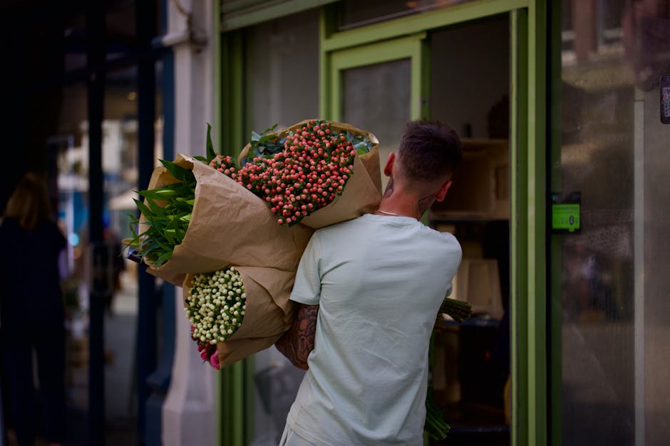 A young man with short brown hair, wearing a light grey T-shirt and visible tattoos on his right arm, is carrying a large bouquet of fresh flowers. The bouquet includes pink and white hypericum berrie