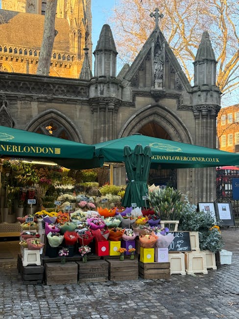 An outdoor flower stand positioned in front of a historic stone church with gothic architecture, featuring pointed arches, spires, and detailed carvings. The stand displays a vibrant array of fresh fl
