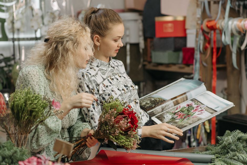Two women in a florist workshop examining a bouquet of red and pink flowers with green foliage, surrounded by various floral materials, wrapping paper, and ribbons. The woman on the left, with curly b