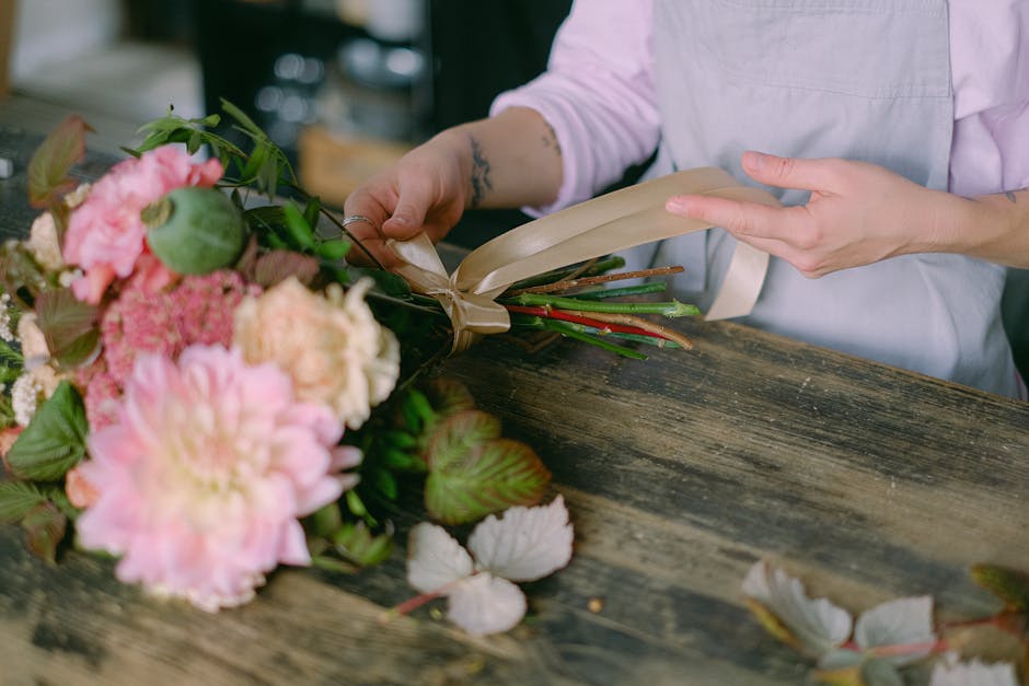 A florist, dressed in a light pink shirt and white apron, is seen preparing a vibrant bouquet of fresh flowers on a rustic wooden table. The bouquet includes large, blooming pink and cream peonies, ac