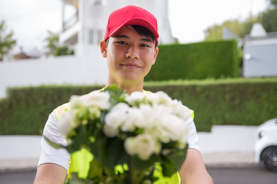 A delivery person wearing a red cap and a high-visibility yellow vest is holding a bouquet of fresh flowers, including white roses and green foliage, in front of him. The flowers are arranged in a com
