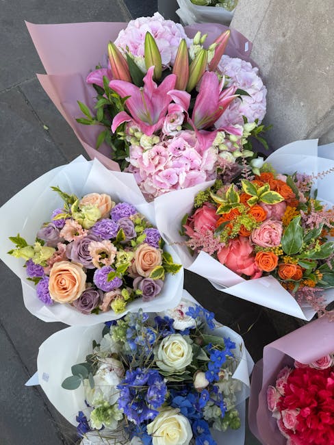 A collection of four floral arrangements and bouquets displayed outdoors on a grey surface, featuring a variety of fresh flowers. The centerpiece bouquet includes large pink lilies, pink hydrangeas, a