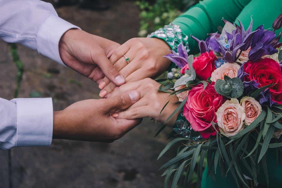 A close-up view of a floral arrangement being handed over, featuring a bouquet composed of vibrant pink roses, soft peach roses, purple irises, and sprigs of eucalyptus with rounded leaves. The bouque