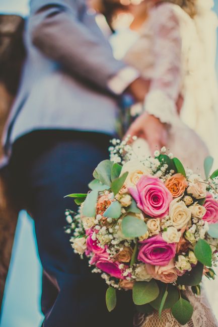 A close-up of a vibrant floral bouquet featuring a mix of pink roses, peach roses, white roses, and sprigs of baby's breath, complemented by green eucalyptus leaves. The bouquet is arranged in a lush,