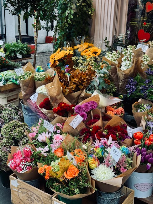 An outdoor flower display featuring a variety of fresh floral arrangements and bouquets wrapped in brown kraft paper, arranged on wooden crates and metal buckets. The flowers include vibrant pink lili