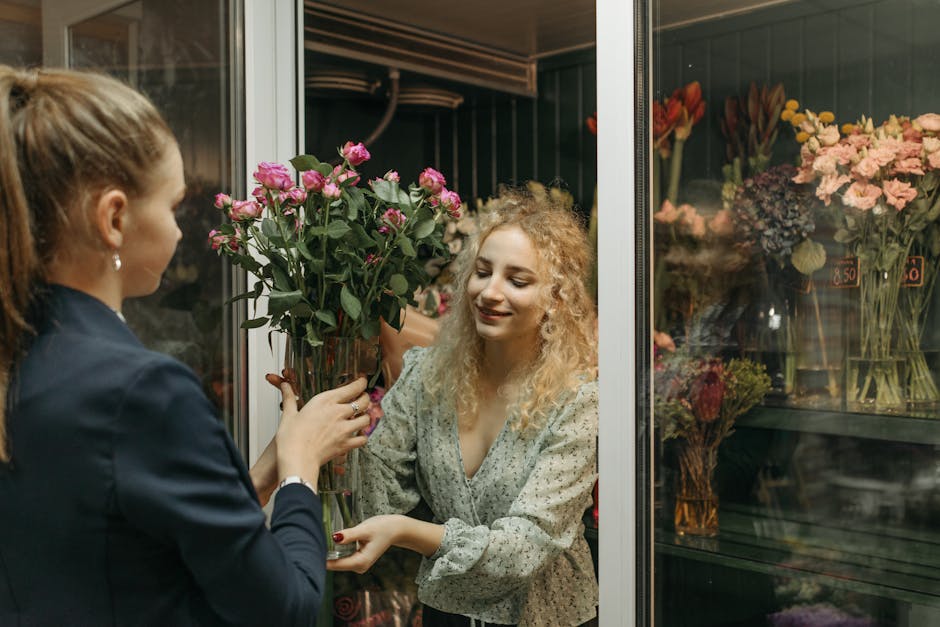 A florist with light skin and long hair tied back is handing a tall, clear glass vase filled with pink roses to a customer with curly blonde hair and light skin, smiling gently. The scene takes place 